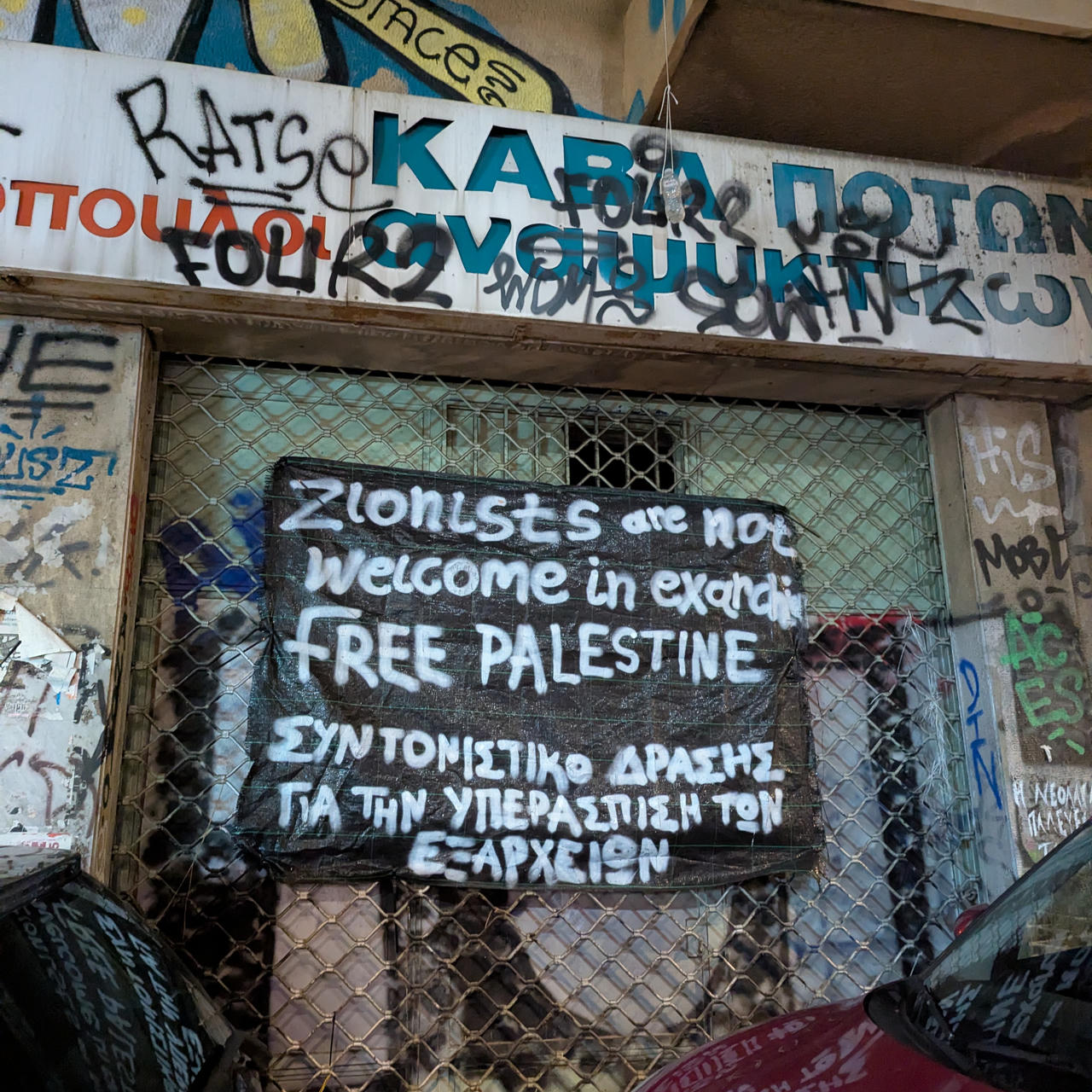 Photo d'une devanture de magasin recouverte de tags, dont un en anglais qui dit "Zionists are not welcome in Exarcheia" (les sionistes ne sont pas les bienvenus à Exarcheia)